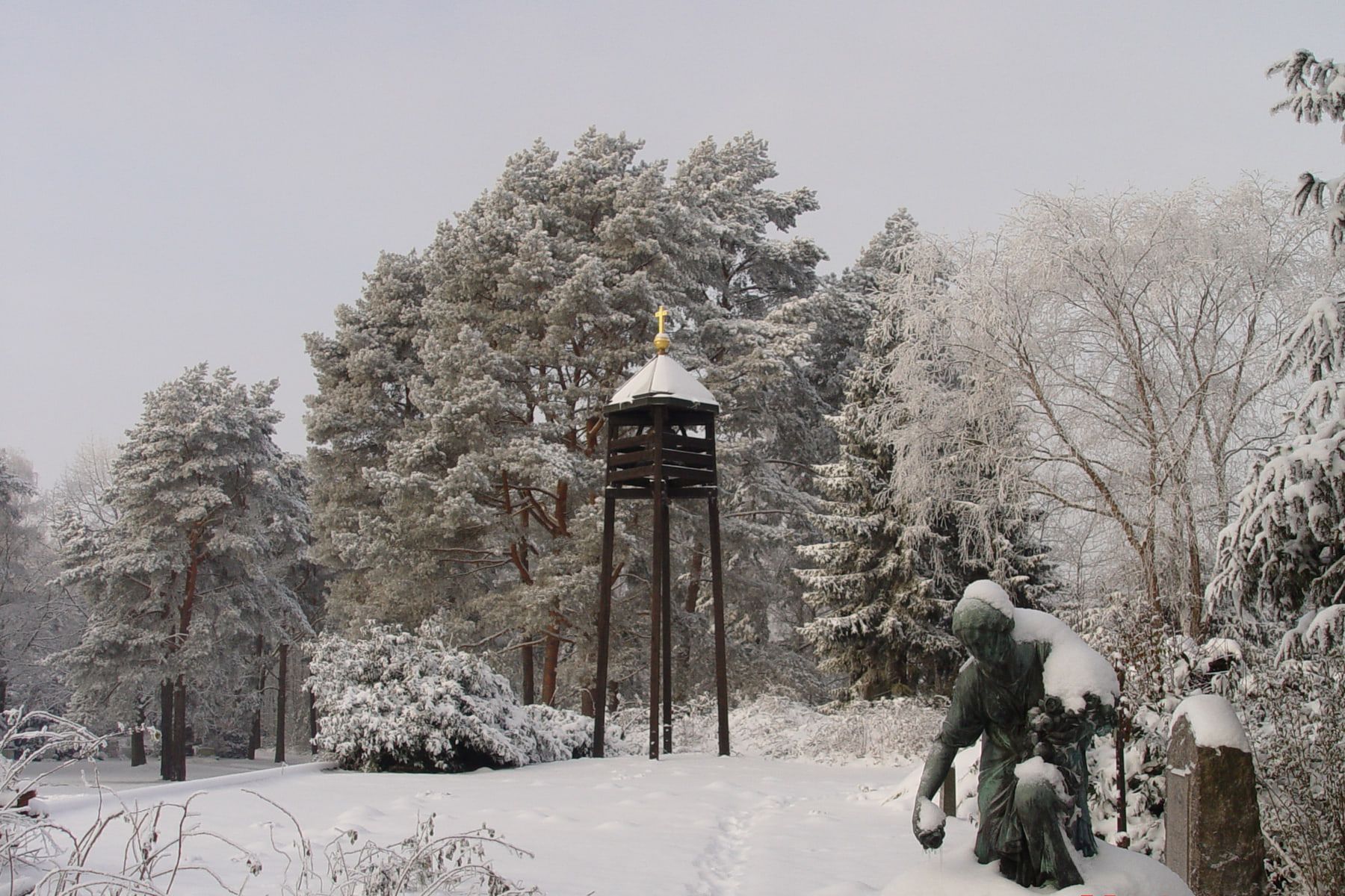 Verschneite Gräber und freistehender dunkler Glockenturm zwischen winterlichen Bäumen auf Friedhof Bergedorf